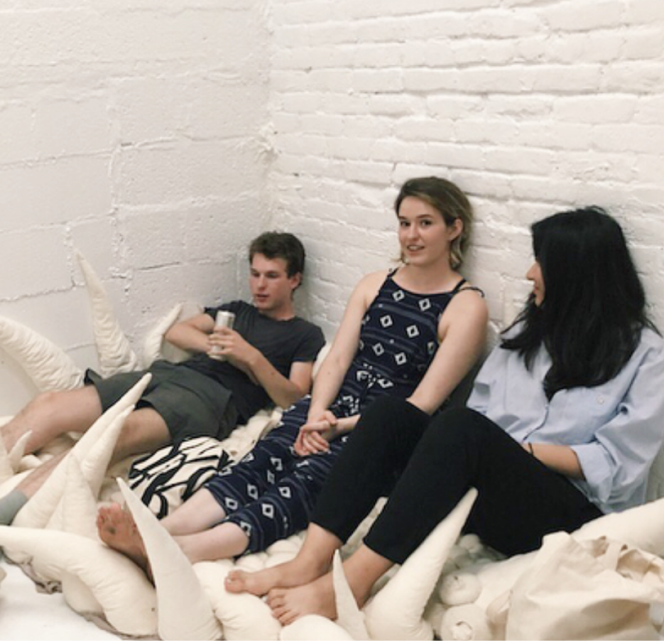 image of three people sitting on a soft, white textile sculpture in the corner of a white gallery. one person is looking at the camera while the other two are in conservation.